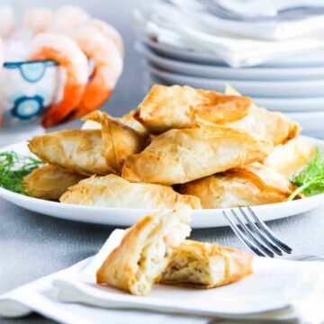 A plate stacked with air fryer shrimp and artichoke phyllo triangles next to a bowl of shrimp cocktail.