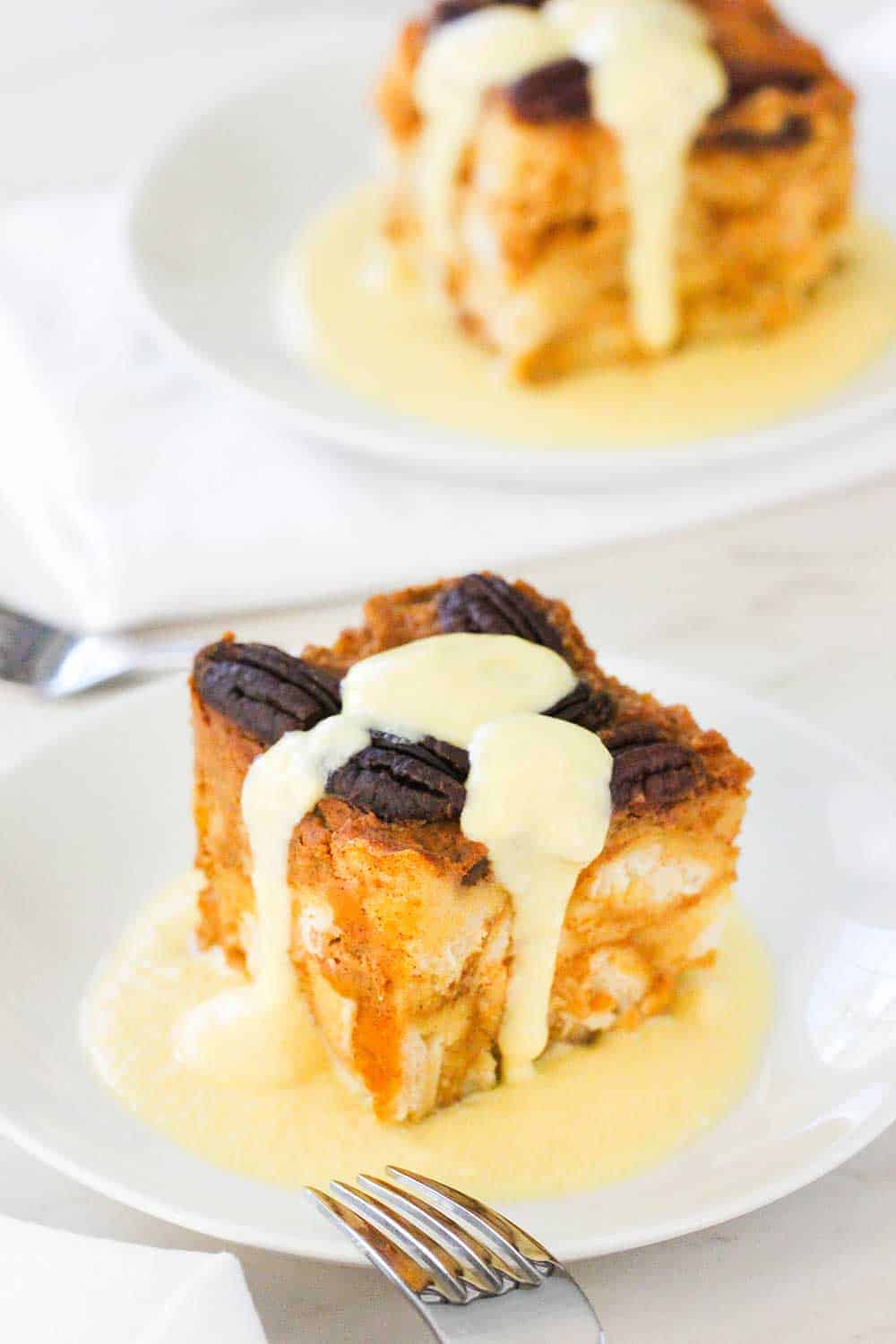 A piece of pumpkin bread pudding with vanilla custard poured over the top on a white plate. 