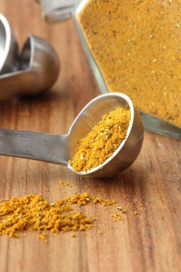 A silver measuring spoon filled with homemade curry powder next to a container of the spice.