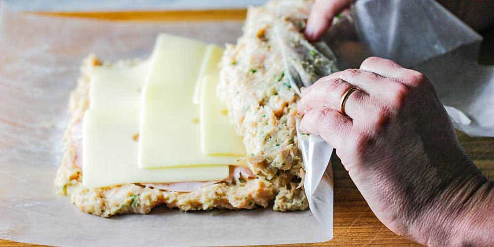 Two hands rolling wax paper filled with cordon bleu meatloaf mixture into a loaf. 
