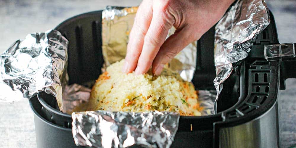 A hand pressing breadcrumbs onto a partially cooked meatloaf in an air fryer. 