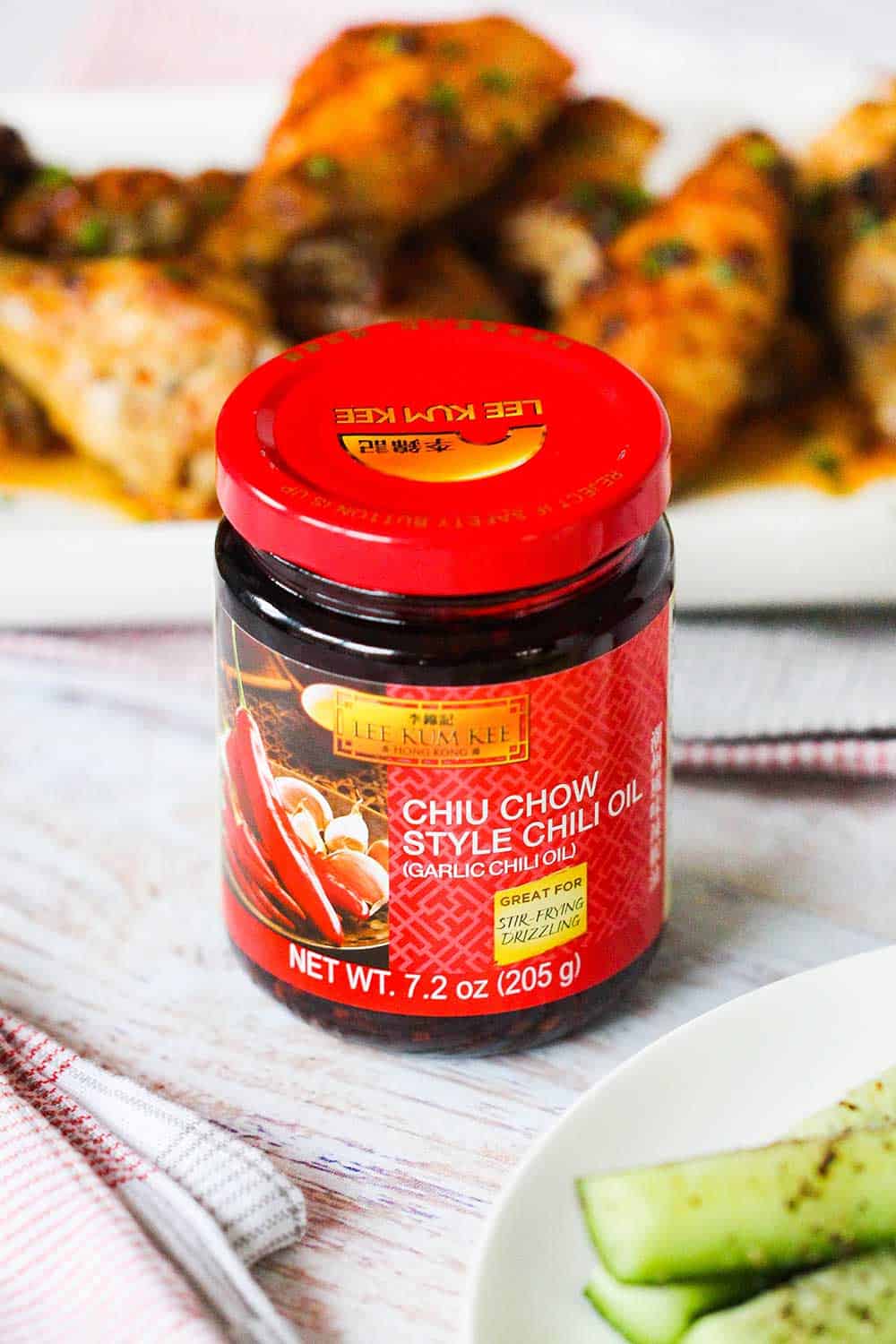 A jar of chili garlic sauce in front of a white platter of baked hot wings and next to sliced cucumbers. 