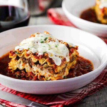 A white bowl of classic meat lasagna next to a glass of red wine.