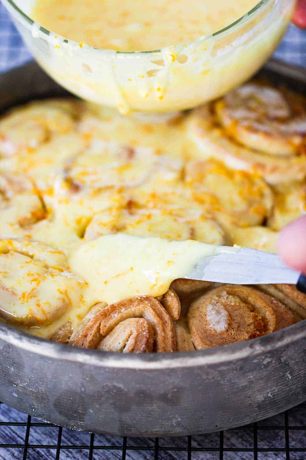 A hand using a small spatula to spread orange icing over baked sweet rolls.