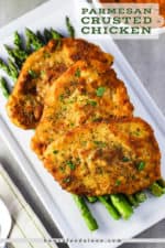 A white serving platter of roasted asparagus with three Parmesan crusted chicken cutlets on top.