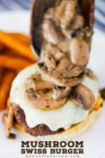 A large wooden spoon placing mushroom sauce onto a mushroom Swiss burger on a plate next to a pile of French fries.