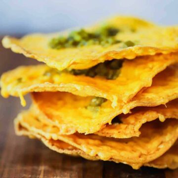 A stack of thin classic nachos topped with jalapeno relish on a wooden cutting board.
