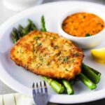 A white dinner plate filled with roasted asparagus topped with Parmesan crusted chicken with a small bowl of vodka sauce next to it.