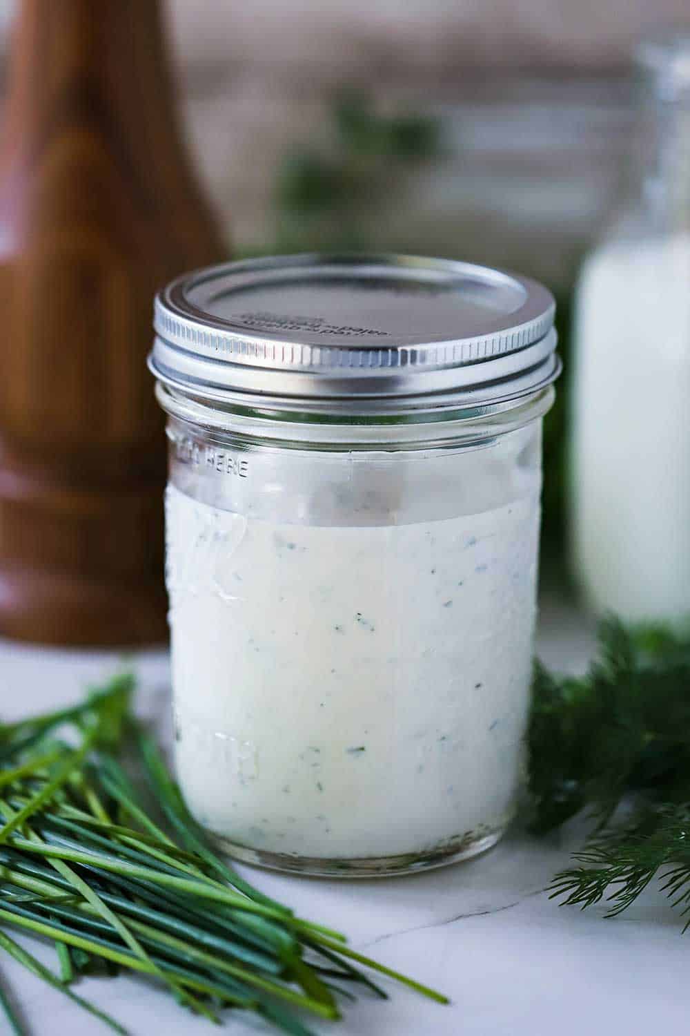 A Mason jar filled with homemade ranch dressing with a lid on top, next to chives, buttermilk, and a pepper grinder. 