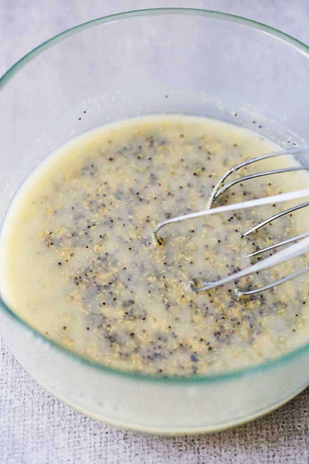 A whisk in a glass bowl holding melted butter with poppy seeds.