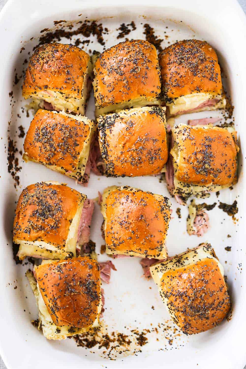 An overhead view of a white baking dish filled with ham and Swiss sliders with one missing.