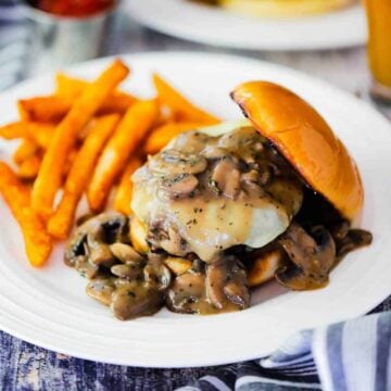A mushroom Swiss burger on a white plate next to a small bowl of ketchup and and a beer.
