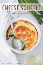 An overhead view of a cheese soufflé with asparagus with a portion missing and a large spoon in the dish and fresh asparagus sitting next to it.