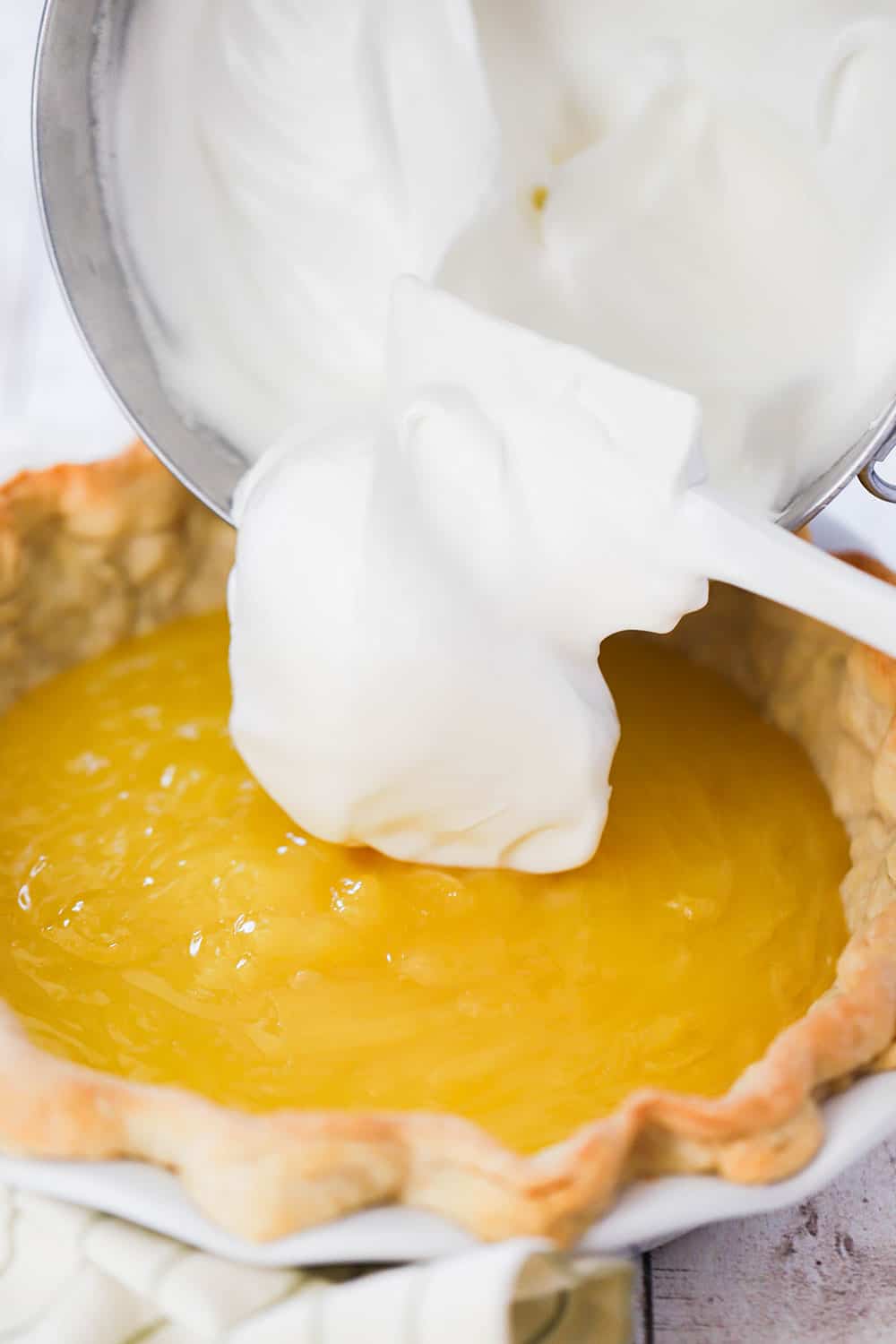 A hand using a white spatula to transfer fluffy meringue onto of lemon curd in a pie shell in a white pie dish.