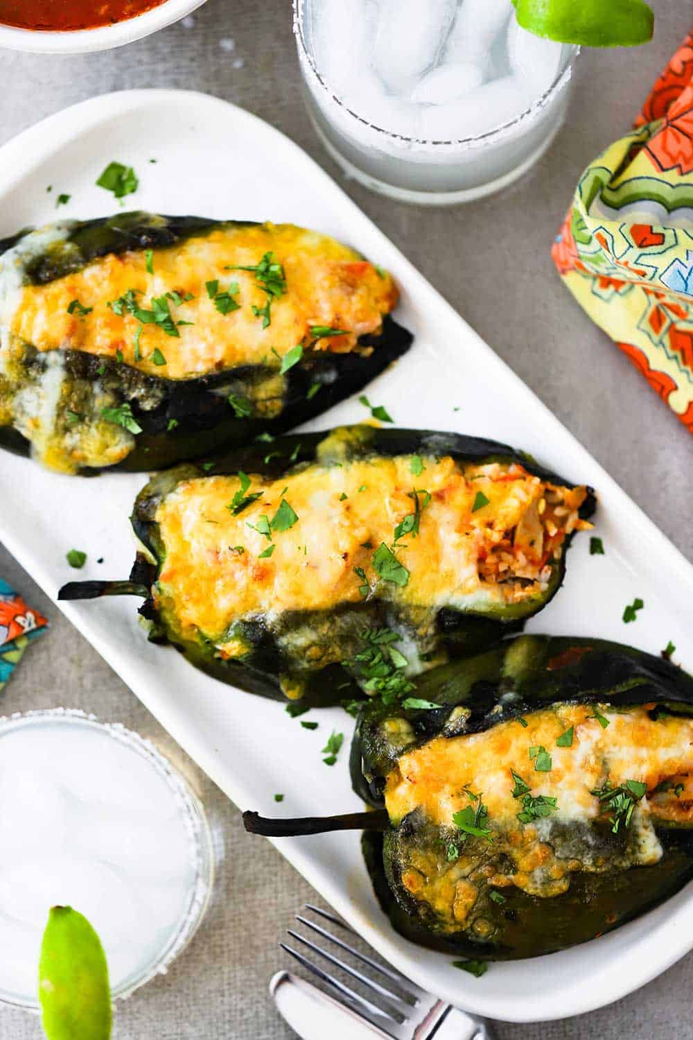 A white platter filled with 3 stuffed poblanos next to 2 margaritas and a festive napkin. 