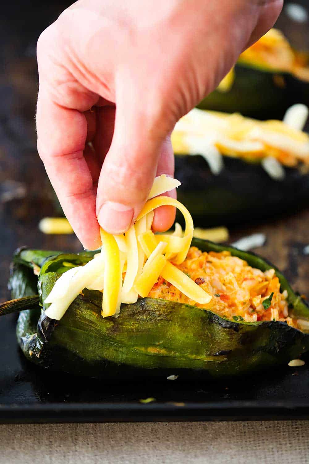 A hand sprinkling shredded cheddar cheese on top of stuffed poblanos on a baking sheet. 
