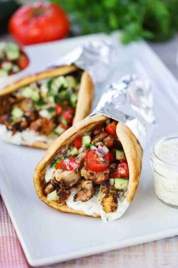 Two chicken gyros wrapped in foil on a white platter next to a small jar filled with tzatziki sauce with a spoon in, with a whole tomato and parsley in the background.