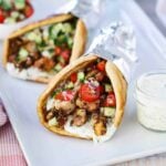 Two chicken gyros wrapped in foil on a white platter next to a small jar filled with tzatziki sauce with a spoon in, with a whole tomato and parsley in the background.