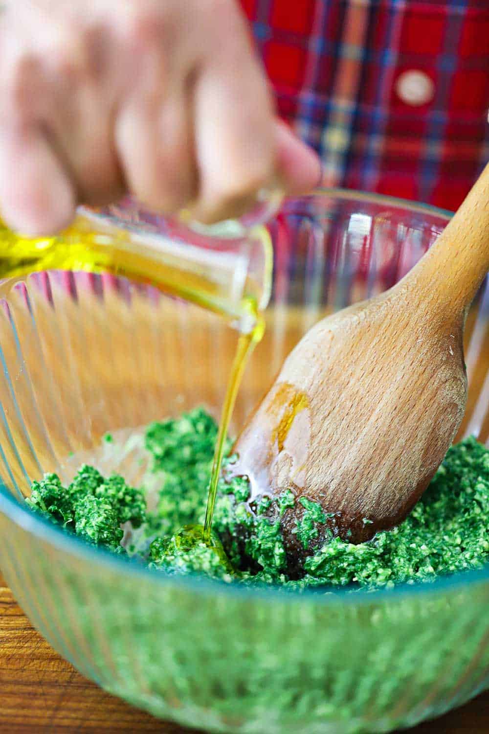 A hand pour olive oil from a glass oil holder into a glass bowl filled with pesto with a large wooden spoon in the bowl.