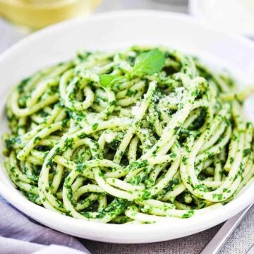 A white bowl filled with pesto pasta next to a grey napkin, two forks, and glass of white wine.