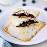 A small white plate holding 2 homemade blueberry pop tarts, one that is split open on top, sitting next to a glass of milk.