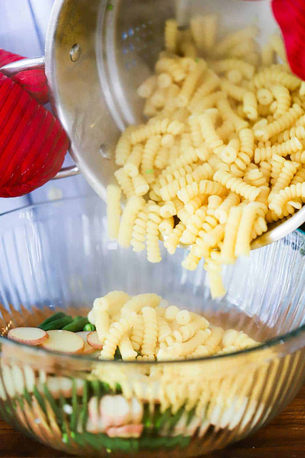 Two hands with red oven mitt on dumping cooked pasta into a glass bowl filled with sliced potatoes and green beans.