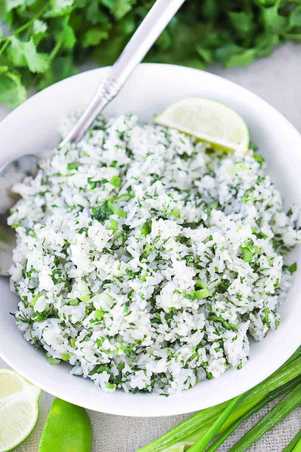 An overhead view of a large white bowl filled with Cilantro Lime Rice next to scallions and a bunch of cilantro, with a silver spoon in the bowl.