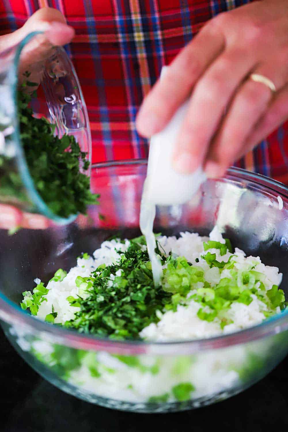 Two hands pouring chopped cilantro and fresh lime juice into a bowl filled with cooked rice and chopped scallions.