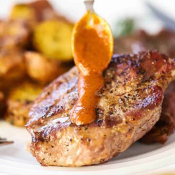 A small spoon drizzling homemade steak sauce down the top and center of a grilled steak on a white plate next to roasted potatoes.