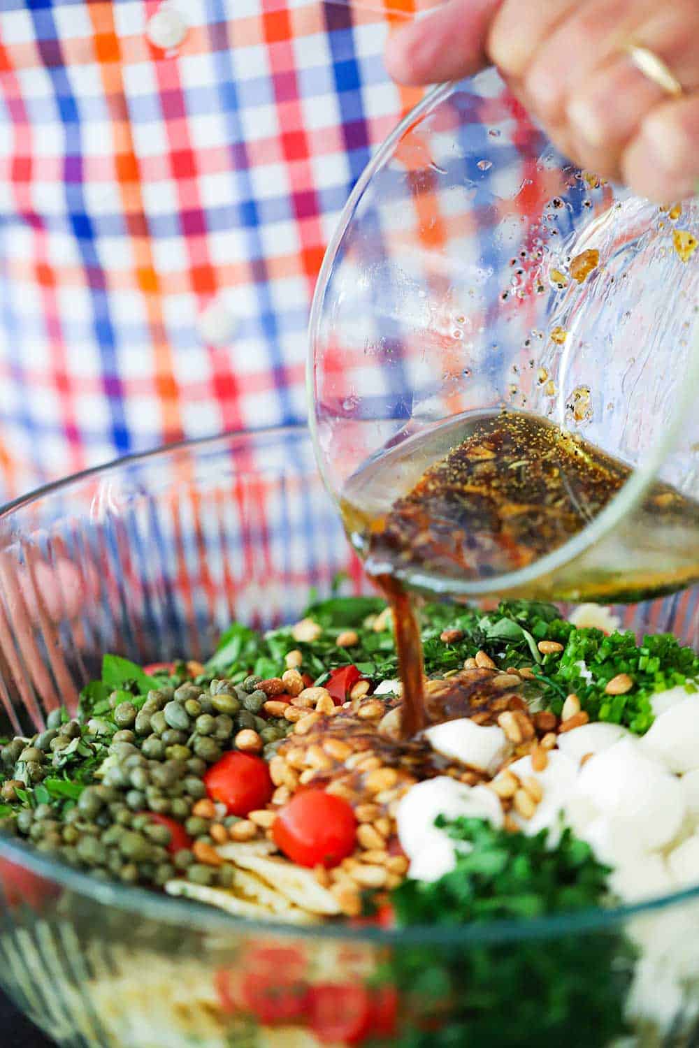 A person pouring a balsamic marinade into a large glass bowl filled with chopped tomatoes, basil, mozzarella, herbs, and pine nuts.
