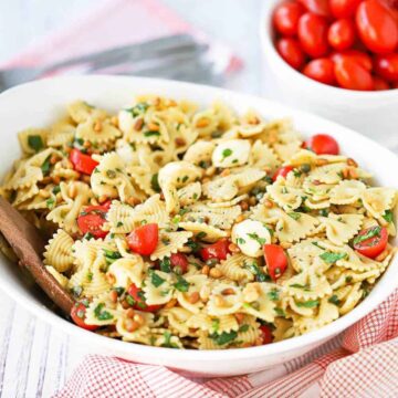 A close-up view of a caprese pasta salad in a large white serving bowl with a wooden spoon in the salad.