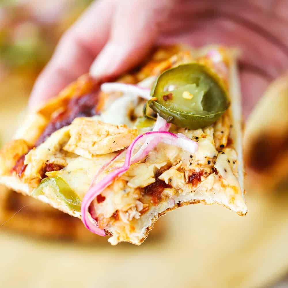 A close up view of a hand holding a slice of BBQ chicken pizza with a bite taken out of it.