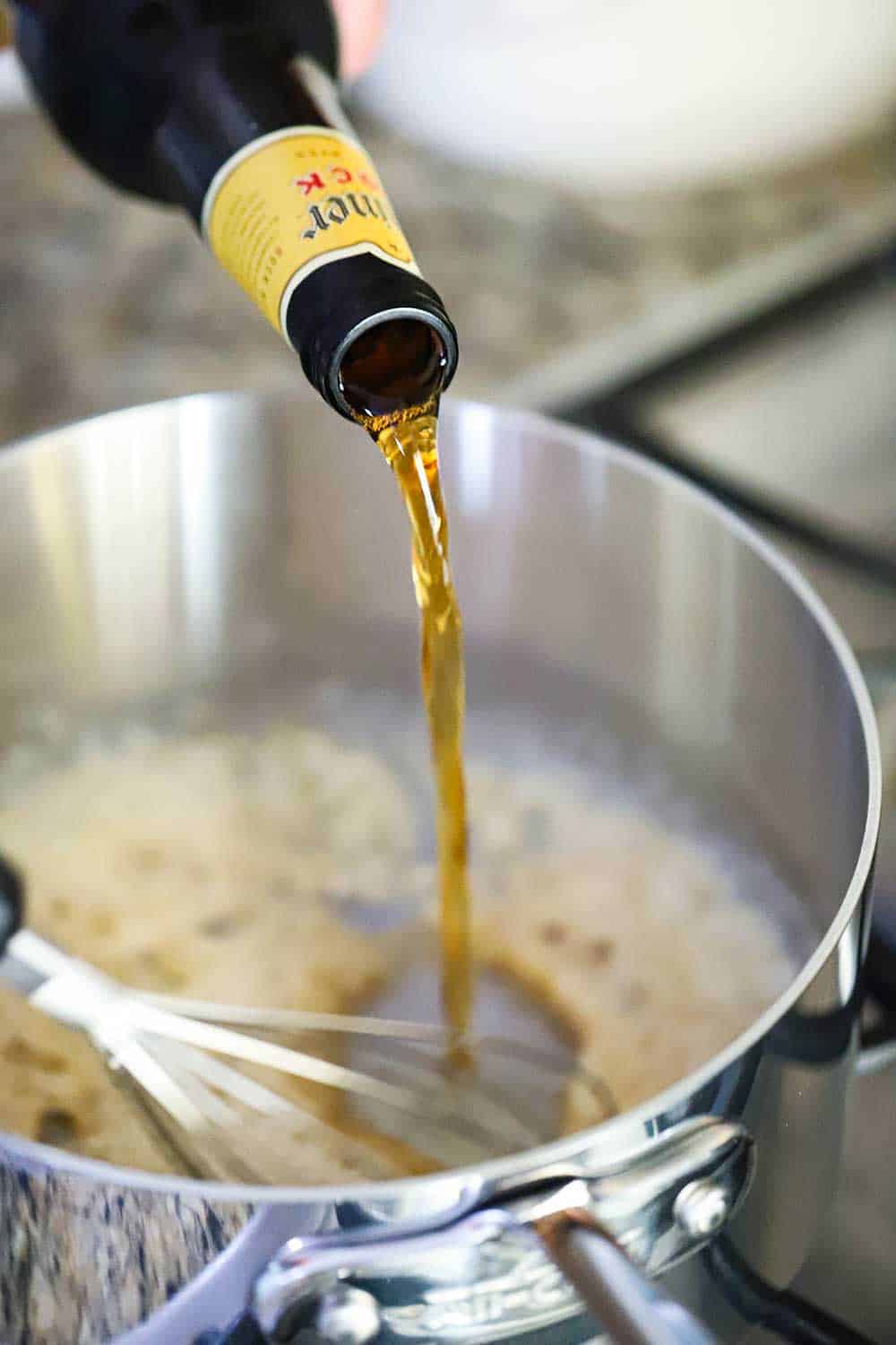 Beer being poured into a stainless still sauce pan with a roux and whisk in it. 