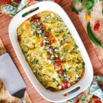 A 9'x13' white baking dish filled with chicken enchiladas with verde sauce topped with chopped tomatoes and cilantro, alongside a metal spatula and festive napkins.