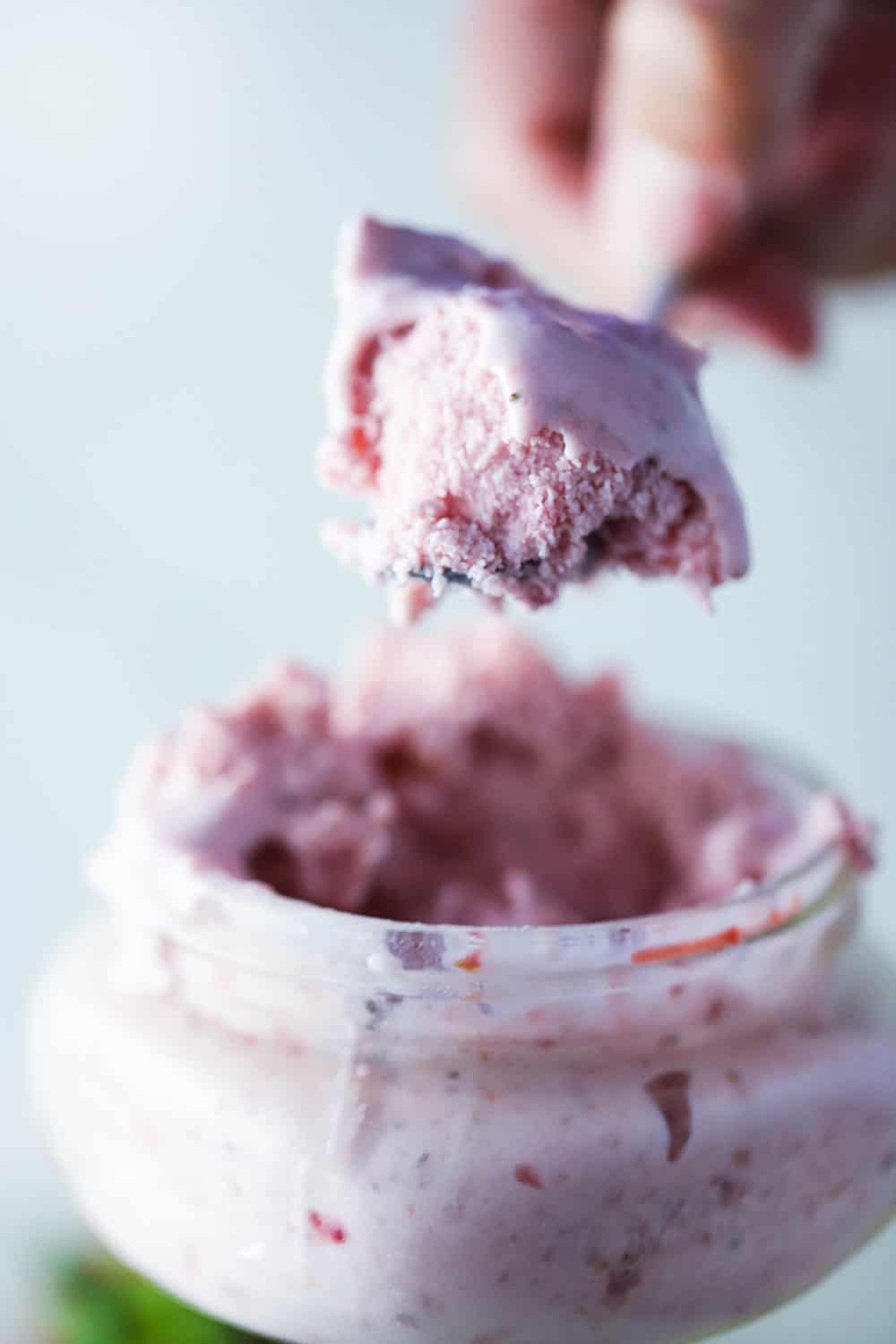 A hand holding up a spoon filled with homemade strawberry ice cream of a glass holder of the ice cream. 