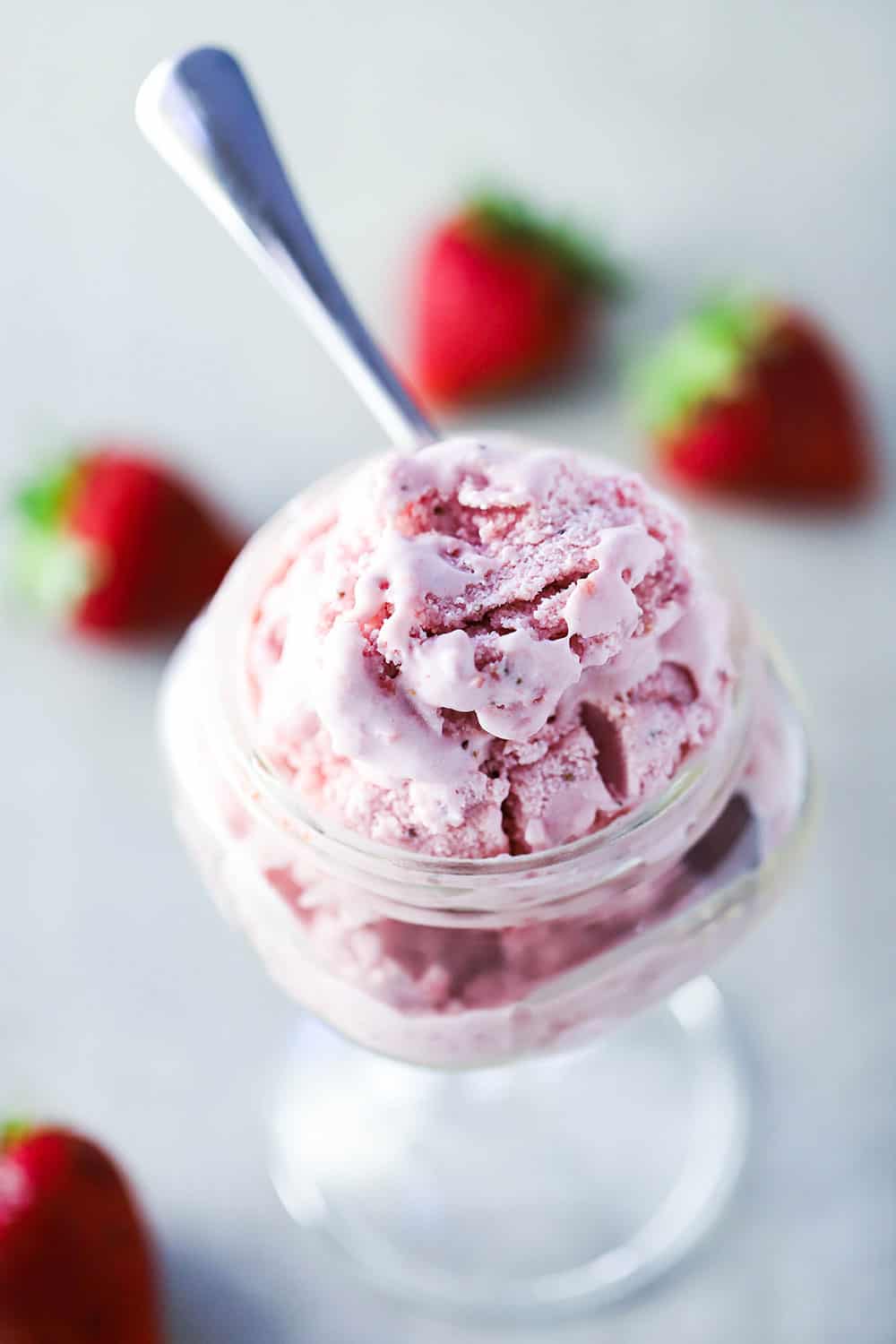 An overhead view of a glass bowl with a stem holding a serving of homemade strawberry ice cream with whole strawberries scattered around next to it. 