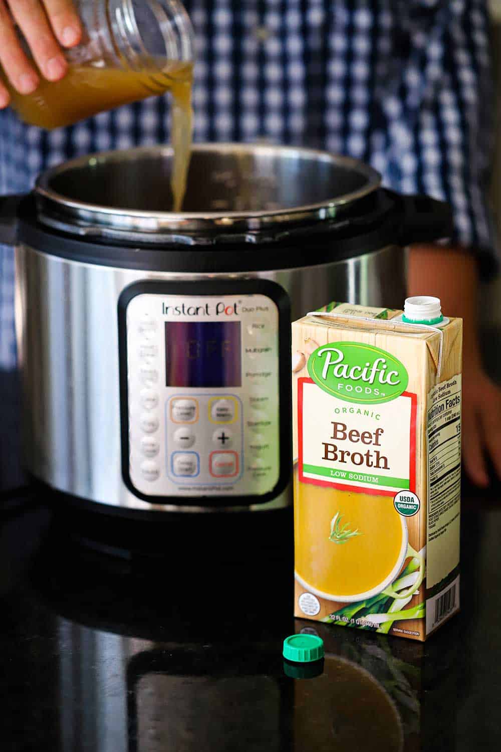 A person pouring beef broth from a jar into an Instant Pot with a box of the broth in front. 