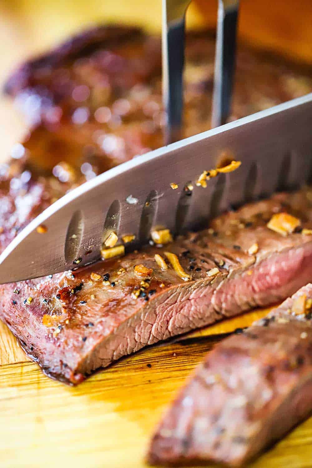 A large chef's knife cutting through a grilled flank steak with a large fork holding it in place. 