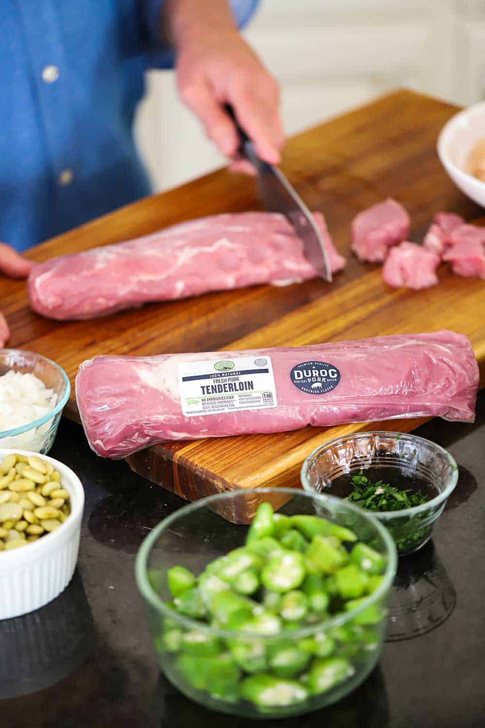 A person cutting bite-sized pieces from a pork tenderloin with a packaged tenderloin in the foreground. 