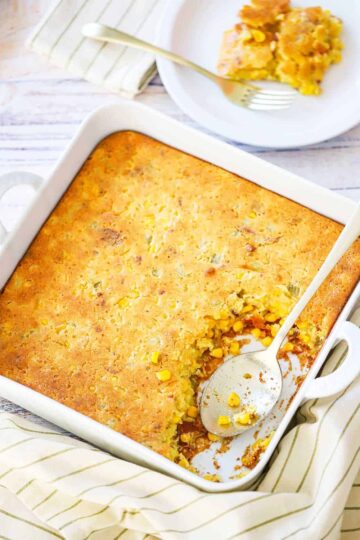 A square white baking dish filled with corn casserole next to a plate filled with the same.