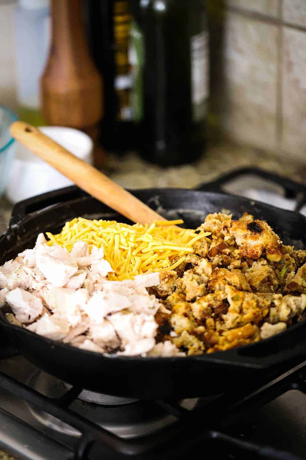 A large black cast-iron skillet filled with roast turkey pieces, broken up dressing, and shredded cheddar cheese, along with a wooden spoon.