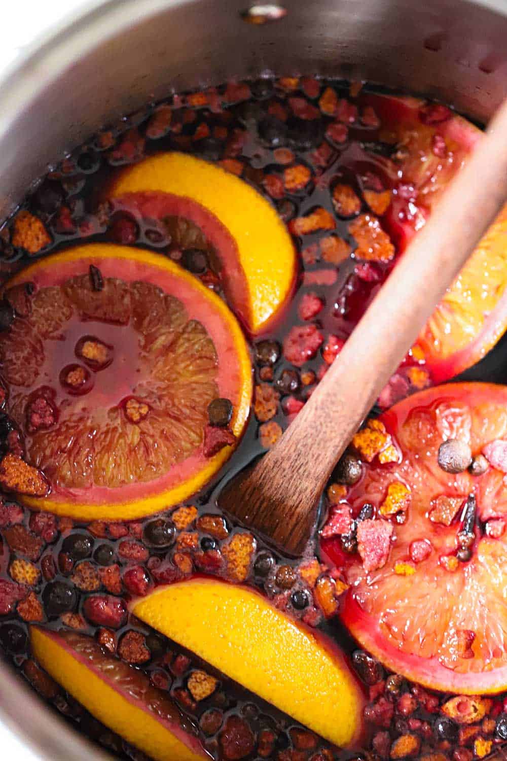 A silver pot filled with red wine, orange slices, and mulling spices with a wooden spoon in the middle of it all.