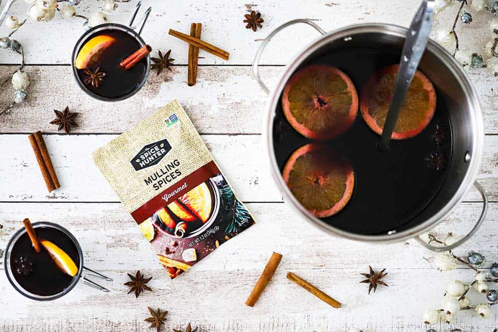A silver pot of mulled red wine with orange slices in it sitting next to a package of mulling spices and two glasses of mulled wine.