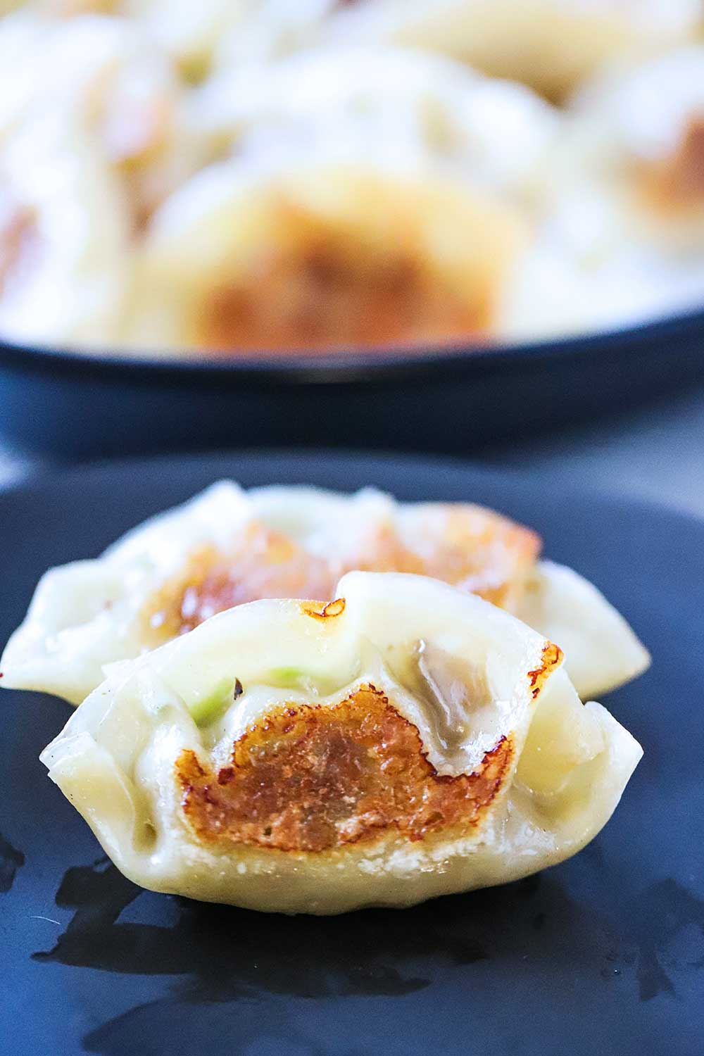 Two cooked potstickers sitting on a black plate with a platter of potstickers in the background.