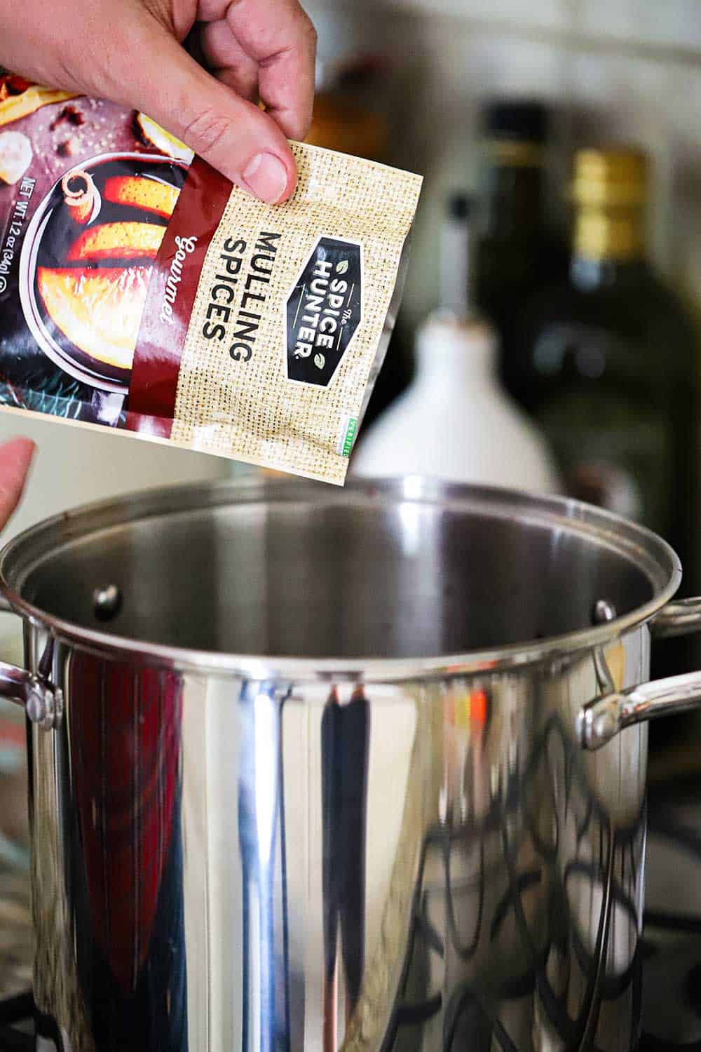 A hand pouring mulling spices from a package into a large silver pot.
