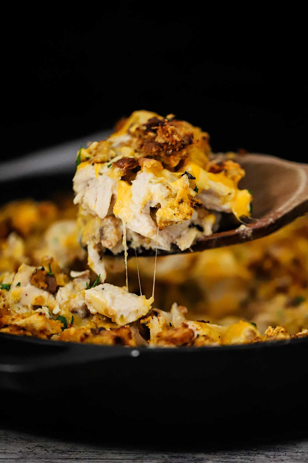A large wooden spoon lifting gooey cheesy turkey and stuffing casserole from a cast-iron skillet.