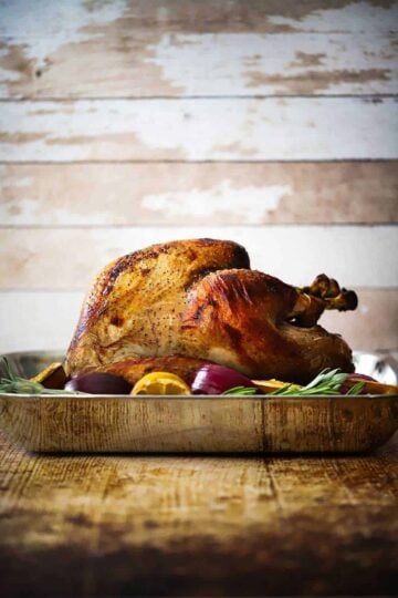 A roasted turkey sitting in a steel roasting pan surrounded by lemons, onions, and herbs.