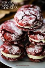 A white plate holding a stack of red velvet sandwich cookies stuffed with cream cheese frosting.