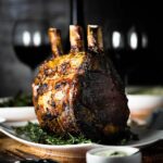 A fully cooked standing prime rib roast sitting upright on a white platter surrounded by fresh herbs.