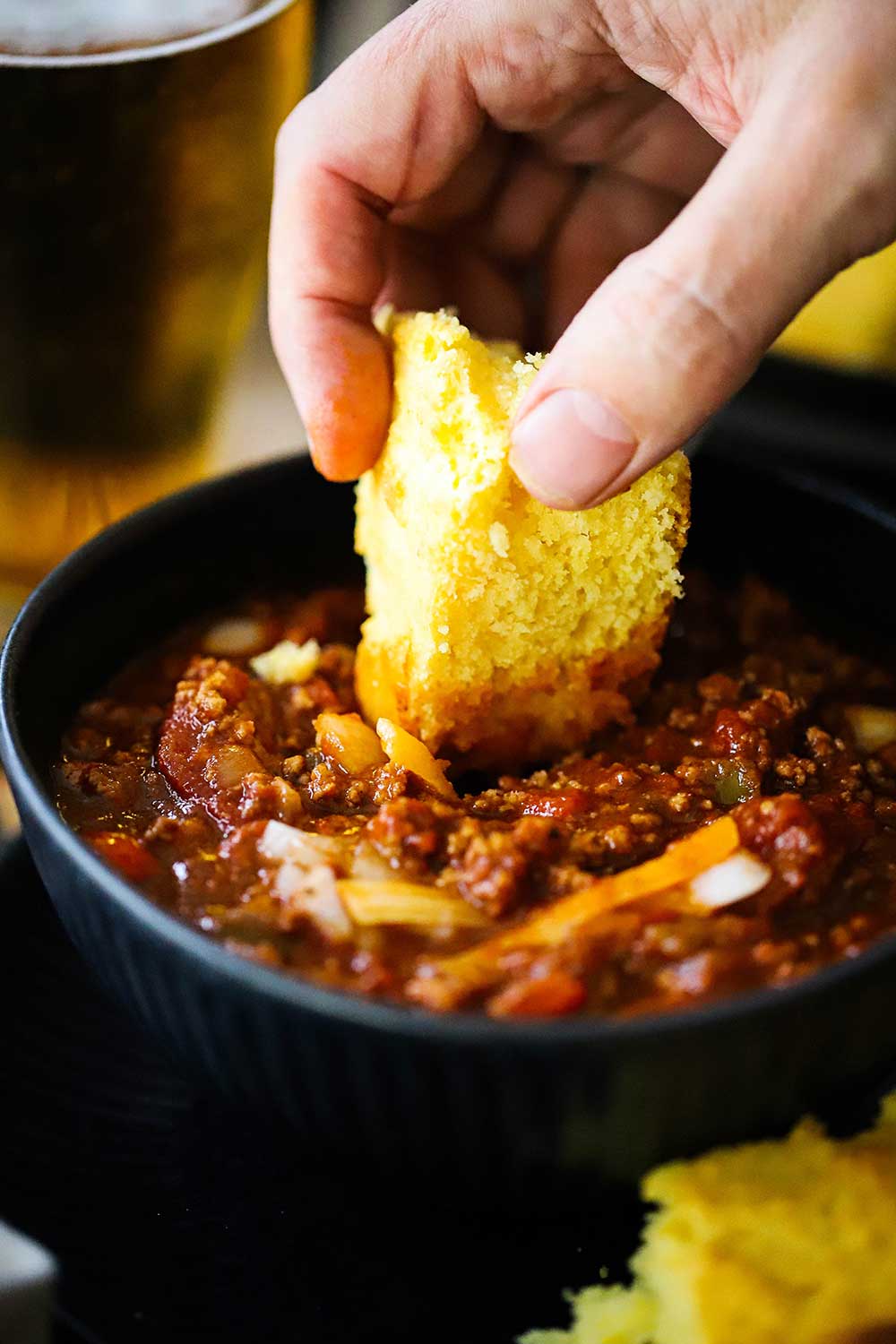 A hand dipping a slice of homemade cornbread into a bowl of turkey chili.